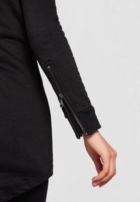 Braccio allungato che indossa un top nero a maniche lunghe con una zip parzialmente aperta vicino al polso su uno sfondo semplice.