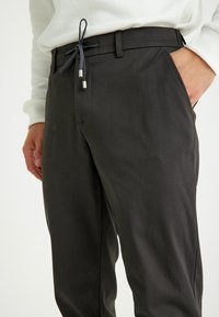 Schwarze, schmal zulaufende Hosen zeichnen sich durch eine glatte Textur, einen elastischen Kordelzug mit silbernen Metallenden, seitliche Taschen und eine schlanke Passform aus.