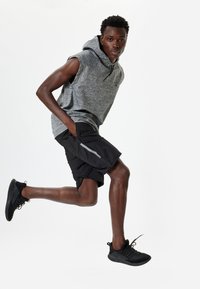 Sudadera sin mangas gris, pantalones cortos deportivos negros con acentos reflectantes y zapatillas de correr negras. Modelo en una pose dinámica de carrera.