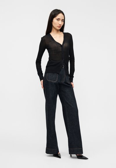 Μαύρη διάφανη ζακέτα με ντεκολτέ σε σχήμα V και κουμπιά μπροστά, σε συνδυασμό με φαρδιά μπλε τζιν παντελόνια με εμφανείς ραφές και πλαϊνές τσέπες.