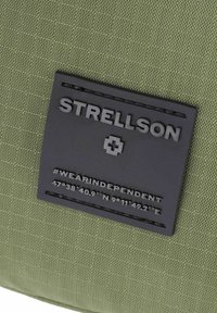 Olivovo zelená textúrovaná tkanina s matne čiernym obdĺžnikovým panelom, na ktorom je napísané "STRELLSON," znak plus a súradnice #WEARINDPENDENT.