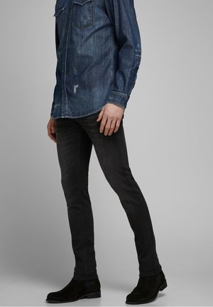 Mann trägt dunkle Slim-Fit-Jeans, schwarze Knöchelstiefel und ein Denim-Hemd mit langen Ärmeln vor schlichtem Hintergrund.