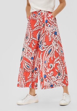 Szerokie spodnie z żywym, abstrakcyjnym kwiatowym wzorem w kolorach czerwonym, białym, niebieskim i zielonym, zestawione z białymi sportowymi butami i rękami w kieszeniach.