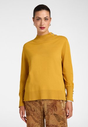 Donna che indossa un maglione giallo senape con dettagli a bottoni sulla spalla e sui polsini, abbinato a pantaloni di velluto marrone con motivo.