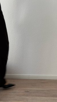 Osoba v černých kalhotách a botách kráčí kolem bílé texturované zdi a světlé dřevěné podlahy, oříznuté z levé strany.