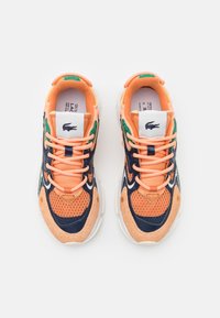 La chaussure présente une tige en mesh pêche avec des accents navy, des détails verts et des lacets orange. Semelle blanche épaisse avec des motifs texturés.