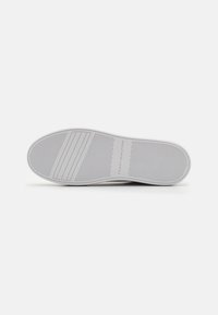 Semelle en caoutchouc blanc d'une sneaker, présentant des rainures horizontales pour l'adhérence et un logo en relief. Design épuré et minimaliste avec une texture lisse.