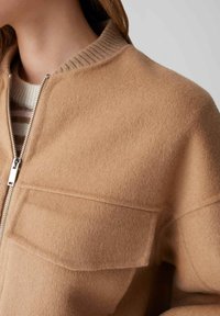 Giacca in misto lana marrone con collo a coste, chiusura con zip e una tasca frontale singola sul lato sinistro. Tessuto morbido e strutturato.