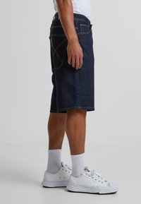 Shorts en denim bleu marine avec une coupe classique, coutures visibles et deux poches arrière. Assorties avec des baskets blanches et des chaussettes côtelées à la cheville.