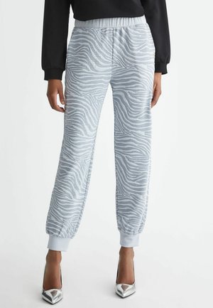 Kvinde iført lysegrå joggebukser med zebra-print, elastiske manchetter og linning, kombineret med sølvfarvede høje hæle og en sort langærmet top.