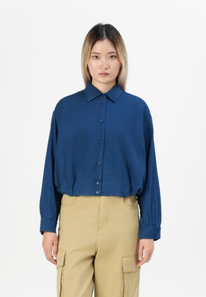 Giovane donna che indossa una camicia lunga a maniche lunghe di colore blu scuro, ampia e con bottoni, e pantaloni cargo beige, in piedi davanti a uno sfondo bianco uniforme.