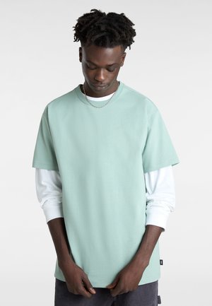 Giovane uomo con pelle scura che indossa una camicia manica corta verde menta a strati e una camicia manica lunga bianca, che guarda in basso con le mani vicino alla vita.