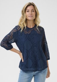 Blusa azul marino con encaje superpuesto que presenta patrones circulares. Las mangas 3/4, el dobladillo festoneado y la tela texturizada realzan el diseño.