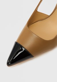 Brązowy skórzany but na wysokim obcasie z zaostrzonym noskiem, z czarną lakierowaną skórzaną nasadką na czubku. Otwarta boki o gładkiej fakturze.