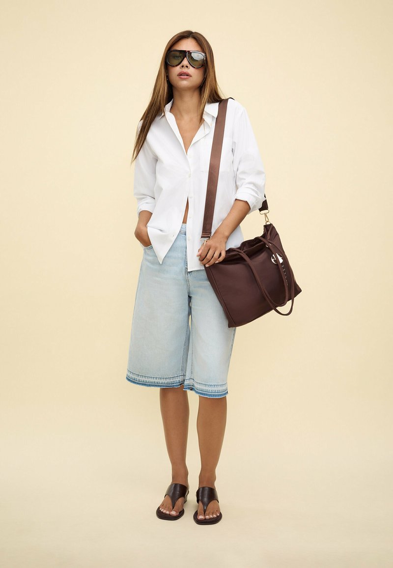 Chemise blanche à bouton, culottes en denim bleu clair avec des détails en bas, sac à bandoulière marron et sandales noires. Grande paire de lunettes de soleil noires.