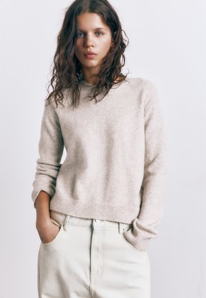 Jeune femme aux cheveux ondulés portant un pull beige et un pantalon clair, debout avec les mains dans les poches devant un fond uni.