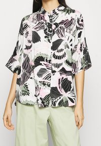 Blouse à manches courtes avec un motif floral en noir, rose et vert sur fond blanc ; présente un devant à boutons et un col.