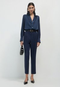 Donna che indossa una blusa di satin blu navy infilata in pantaloni tailleur blu navy con cintura nera, mentre tiene una borsa nera e indossa tacchi a punta neri.