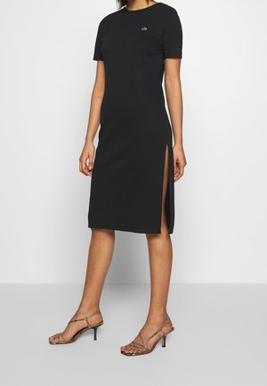 Femme portant une robe midi noire à manches courtes avec une fente latérale et un petit logo, assortie à des sandales à talons à lanières.