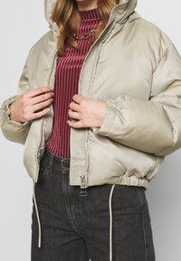 Fyllig beige jacka med huva, dragkedjestängning, elastiska ärmar och dragsko i fållen, bärs över en ribbad vinröd topp och mörka jeans.