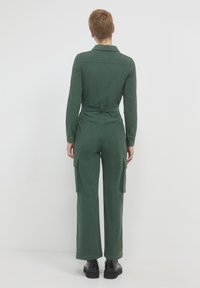 Salopette en velours côtelé vert avec col, manches longues, taille ceinturée et jambes évasées ; comporte des poches cargo sur les côtés et des poignets à boutons.