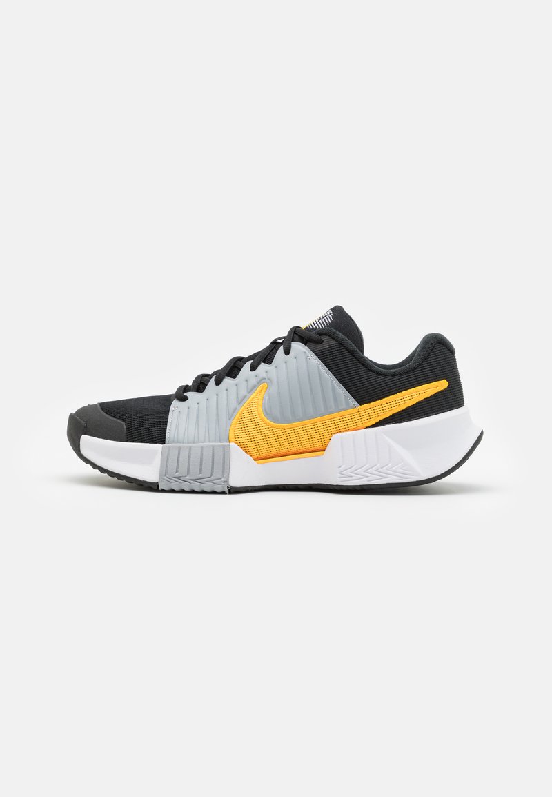 Zwarte en grijze sportieve schoenen met een textuur bovenkant, voorzien van een opvallende oranje swoosh en een witte rubberen zool. Minimalistisch ontwerp.