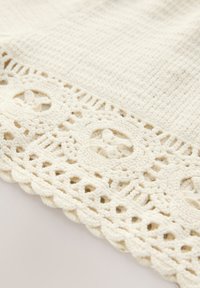 Háčkovaná bílá látka s texturovaným povrchem, zdobená krajkovým okrajem a kruhovými vzory podél spodního okraje.