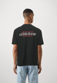 Juodas medvilninis marškinėlis su "GUESS JEANS" logotipu baltos ir raudonos spalvos ant nugaros. Klasikinio stiliaus, trumpomis rankovėmis ir apvaliu iškirpimu.