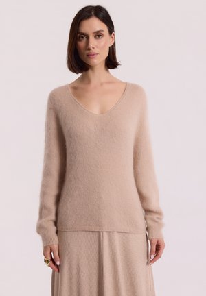 Jasno beżowy sweter z dekoltem w serek, wykonany z miękkiego, puszystego materiału, z długimi rękawami i luźnym krojem. Długość u dołu jest nieco dłuższa z tyłu.