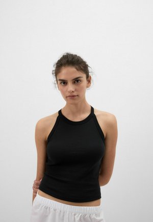 Jonge vrouw met donker haar naar achteren gebonden, draagt een zwarte mouwloze top en witte broek, staand tegen een effen witte achtergrond.