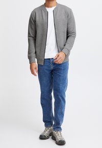 Veste bomber grise texturée avec une fermeture éclair noire, portée sur un t-shirt blanc, associée à un jean bleu et des baskets grises.