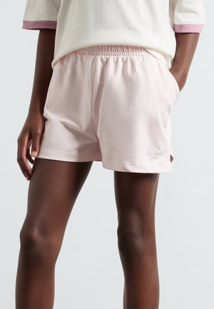 Person trägt hellrosa Shorts mit elastischem Bund und ein weißes Oberteil mit rosa Bündchen, steht mit einer Hand in der Tasche vor einfarbigem Hintergrund.