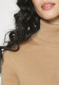 Pull beige à col roulé avec une texture côtelée, vu des épaules jusqu'en haut. Tissu doux avec un design de col ajusté et des détails de couture subtils.