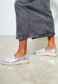 Silberne Slippers mit glänzender Oberfläche, ausgestattet mit einem gepolsterten Obermaterial und einer kontrastierenden weißen Gummisohle. Getragen mit einem Jeansrock.