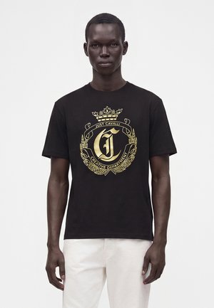 Mand iført en sort t-shirt med et guldvåbenskjold og tekst "Just Cavalli Creative Department" kombineret med hvide bukser, stående mod en hvid baggrund.