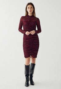 Czerwona i czarna sukienka w panterkę z długimi rękawami, marszczoną fakturą, dopasowanym krojem i długością do połowy ud. Para wysokich czarnych botków.