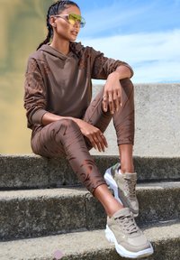 Brauner Hoodie mit orangefarbenen Akzenten, gemusterte braune Leggings und beigefarbene Sneakers mit weißen Sohlen. Modell sitzt im Freien auf Steinstufen.
