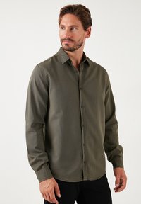 Camisa de botones de manga larga en verde oliva con un tejido texturizado, cuello clásico y botones negros. Ajuste entallado con dobladillo recto.