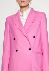 Blazer doppiopetto in tessuto rosa brillante con finitura testurizzata, bottoni neri e colletto a punta. Presenta tasche laterali.