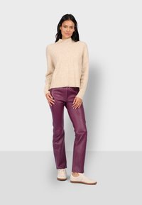 Mujer de pie vestida con un suéter de canalé beige, pantalones de cuero morados y zapatillas blancas contra un fondo gris claro.