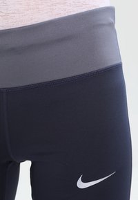 Zbliżenie granatowych legginsów Nike z szerokim szarym pasem w talii i białym logo Nike na udzie.