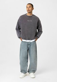 Grijze oversized sweatshirt met geborduurde tekst, gecombineerd met losse blauwe denim jeans en witte sneakers, tegen een effen achtergrond.
