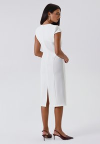 Donna che indossa un abito midi bianco aderente con maniche a cappuccio e spacco posteriore, abbinato a décolleté slingback rosso scuro e orecchini a cerchio dorati.