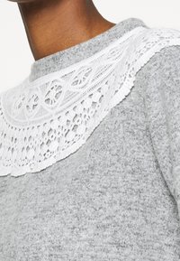 Šedý svetr s bílým krajkovým límcem s složitými vzory a vlnitými okraji, který zdůrazňuje texturovanou tkaninu pro vizuální zajímavost.