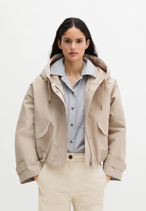 Γυναίκα με μακριά σκούρα μαλλιά που φοράει μπεζ ζακέτα με φερμουάρ και κουκούλα, ανοιχτό μπλε πουκάμισο με κουμπιά και κρεμ παντελόνι ψηλόμεσο, στέκεται με τα χέρια στις τσέπες.
