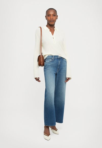 Vrouw in een witte geribbelde lange mouwen polo, blauwe wijde jeans, witte puntige hakken en een bruine schoudertas op een witte achtergrond.