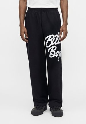 Homme portant un pantalon noir à jambes larges avec le texte blanc « Bill Boys » sur une jambe, une chemise blanche et des baskets noires sur fond blanc.