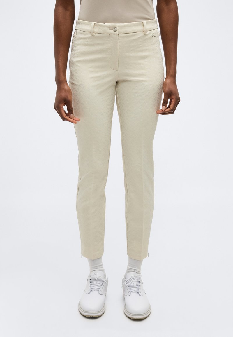 Άτομο που φοράει μπεζ παντελόνι στενής γραμμής με κούμπωμα με κουμπιά, λευκές κάλτσες και λευκά δερμάτινα παπούτσια με κορδόνια, στέκεται μπροστά σε ένα απλό φόντο.