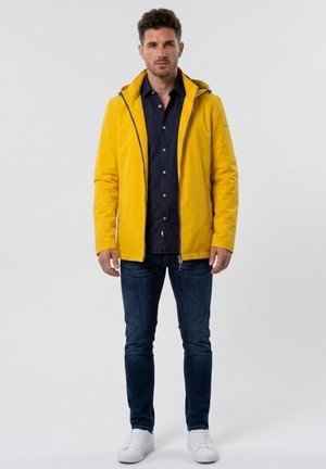 Hombre de pie con una chaqueta amarilla brillante con capucha sobre una camisa de botones azul oscuro, jeans oscuros y zapatillas blancas sobre un fondo liso.