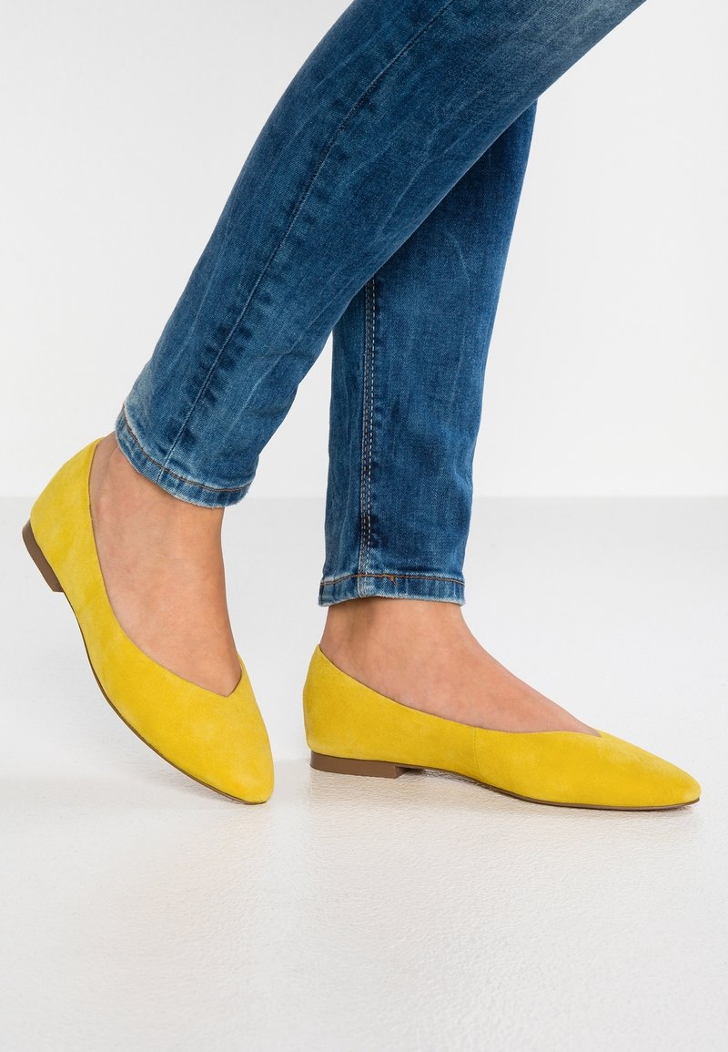 Ballerines en daim jaune avec un bout pointu, un talon plat et une texture lisse ; portées avec un jean bleu ajusté sur fond blanc.